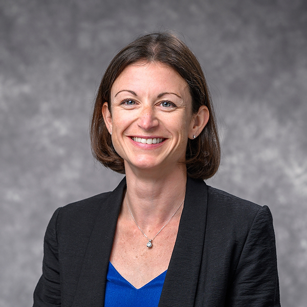 Kelly Tyrrell, sits in a dark blazer and royal blue blouse. She smiles broadly at the camera, her bob length dark hair curling slightly toward her chin.
